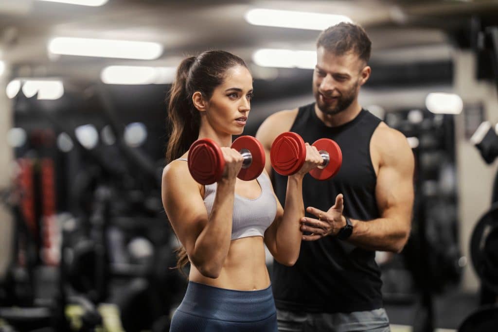 Image shows a man and a woman in a gym. The man appears to be instructing the woman on how to properly lift dumbbells. The woman is holding two, red dumbbells.
