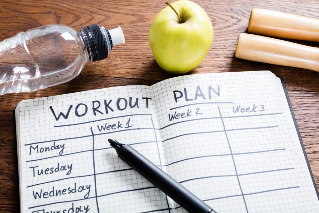 Image shows an overview of a notebook. Written across its white pages are "Workout Plan", followed by the days of the week, and numbers of weeks. There is a black marker on the notebook, as well as a green apple and a water bottle on the table.