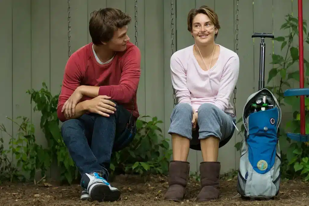 A scene from The Fault in Our Stars showing a young couple sitting on swings, smiling at each other in a park