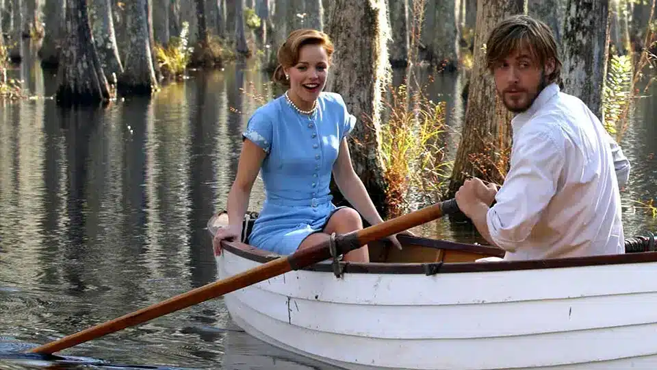 A romantic scene from The Notebook showing a couple in a small rowboat on a serene lake surrounded by trees.