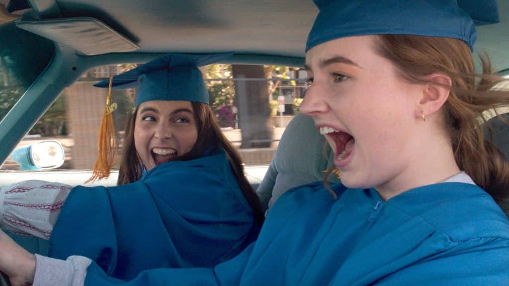Beanie Feldstein and Kaitlyn Dever scream in excitement as they drive off, wearing graduation caps and gowns.