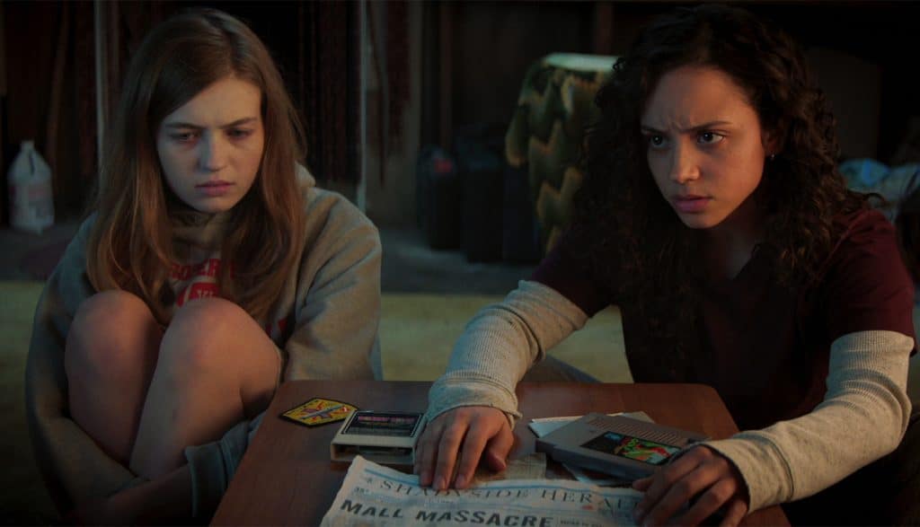Olivia Scott Welch and Kiana Madeira sit side by side on the ground in Fear Street: Part One - 1994. In front of them are papers and clues to the curse of Sarah Fier.
