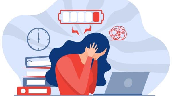 A woman suffering from burnout sits at her desk with her head in her hands, surrounded by a ticking clock with a low battery over her head