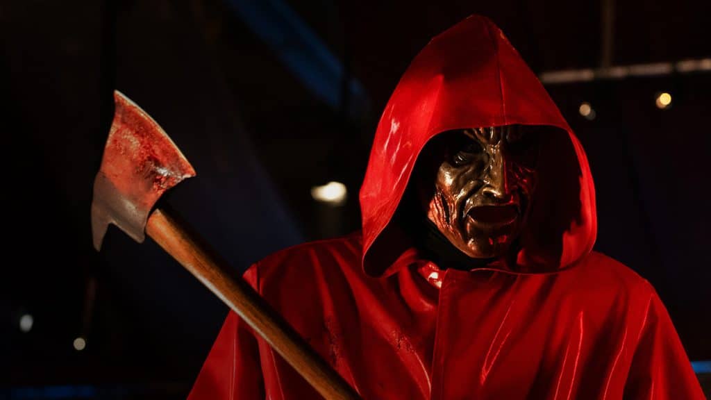 Someone in a red hooded robe, a shiny and exaggerated mask, holding an axe intent on murder.