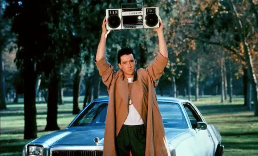 John Cusack as Lloyd Dobler in Say Anything, holding a boombox above his head in a long coat.