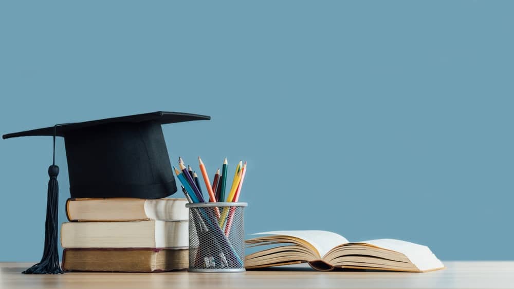 Graduation day. A mortarboard and graduation scroll on stack of books with pencils color in a pencil case on blue background. Education learning concept.