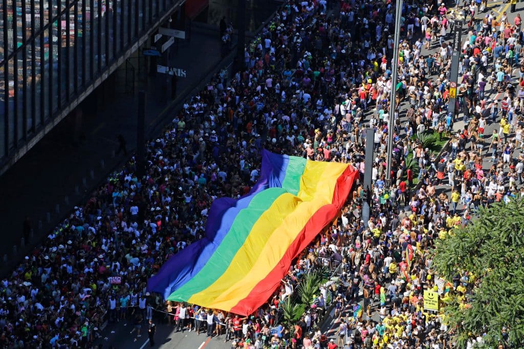 Large crowd marches while carrying a rainbow pride flag.