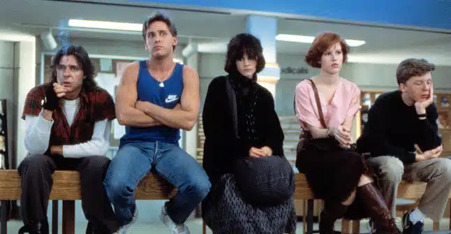 From left to right: Judd Nelson, Emilio Estevez, Ally Sheedy, Molly Ringwald, and Anthony Michael Hall sit on a table in detention, looking annoyed or confrontational.