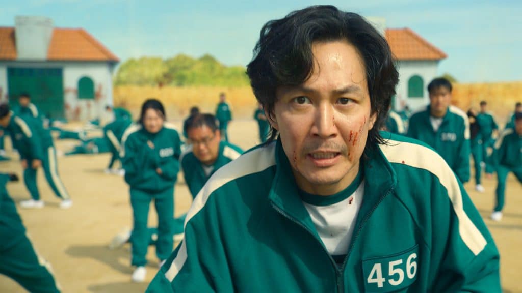 Lee Jung-jae in a green track suit, scared. Behind him, the blurry outlines of other scared individuals in green track suits stand frozen with a blue sky behind them.