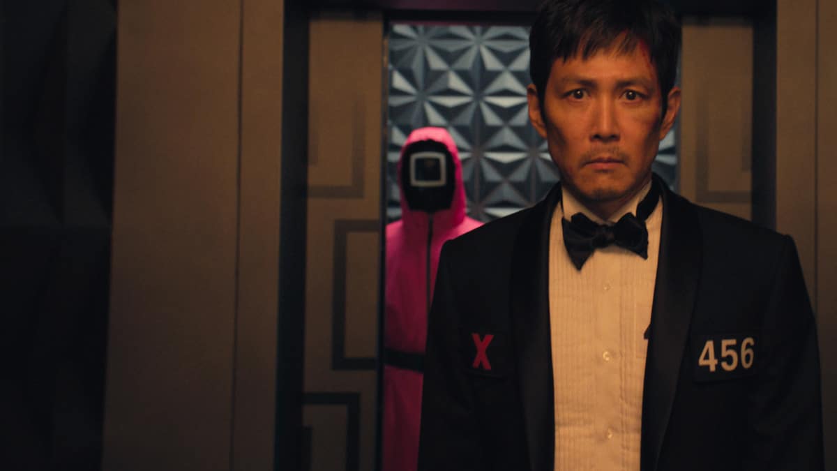 Lee jung-jae stands in a suit, looking past the camera expectantly. Behind him a guard in a mask and pink outfit watches him through a closing door.