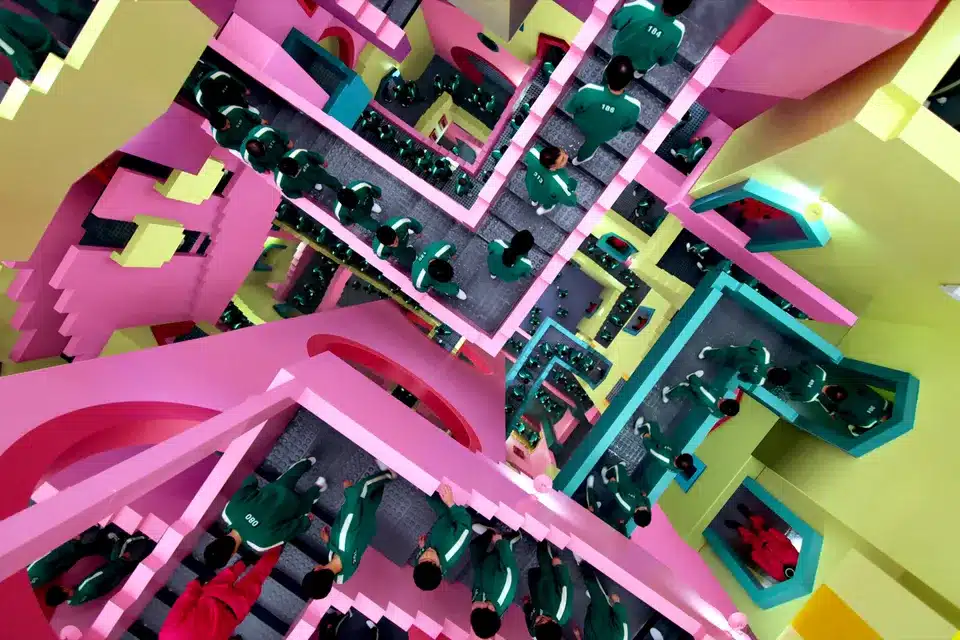 A series of brightly colored and intersecting staircases are shown from above. People in green track suits flood the stairs, guided by guards in bright pink suits and masks.