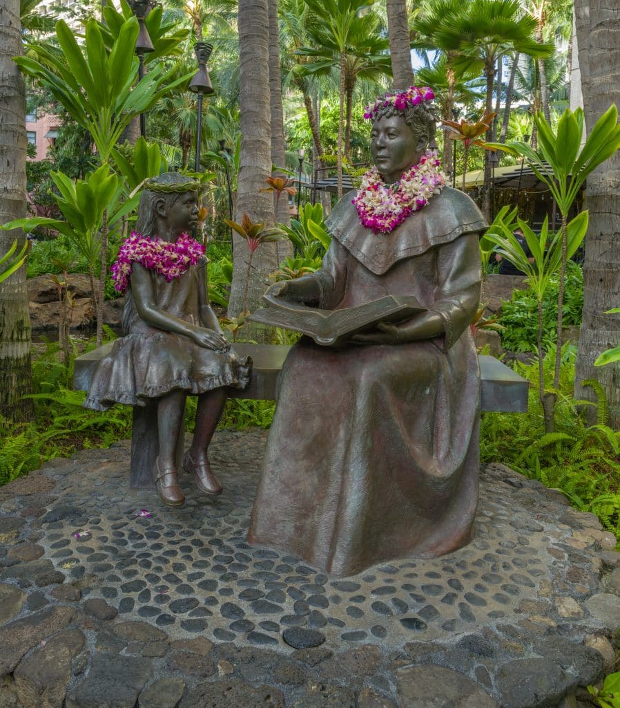 Two statues of a little girl and Queen Liliuokalani sitting while the Queen reads to the girl. Both are decorated in leis