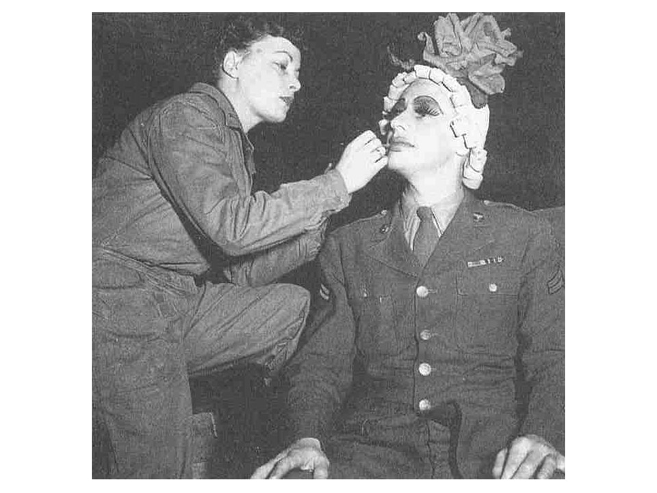 A WAC applies makeup to a solider in an exaggerated, drag style during World War II.