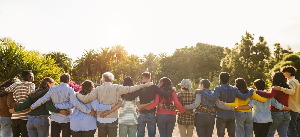 A group of 16 people in a line, their backs towards the camera, have their arms around each other. Change is scary.