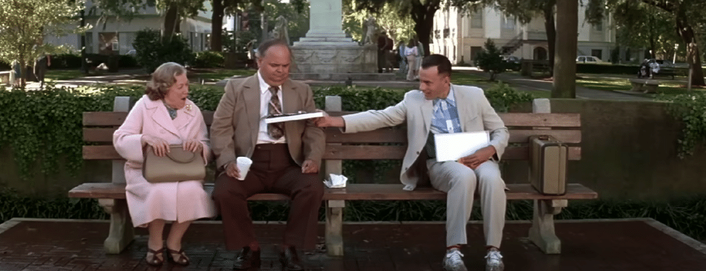 Man sat on bench handing box of chocolates to 2 elderly people.
