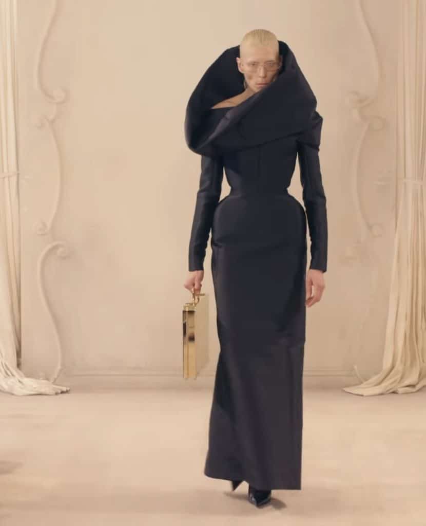 A Balenciaga model walks the runway in black, long sleeve dress which reaches the model’s ankles. The dress also has a large collar which surrounds the model’s face and neck, similar to a turtle neck.
