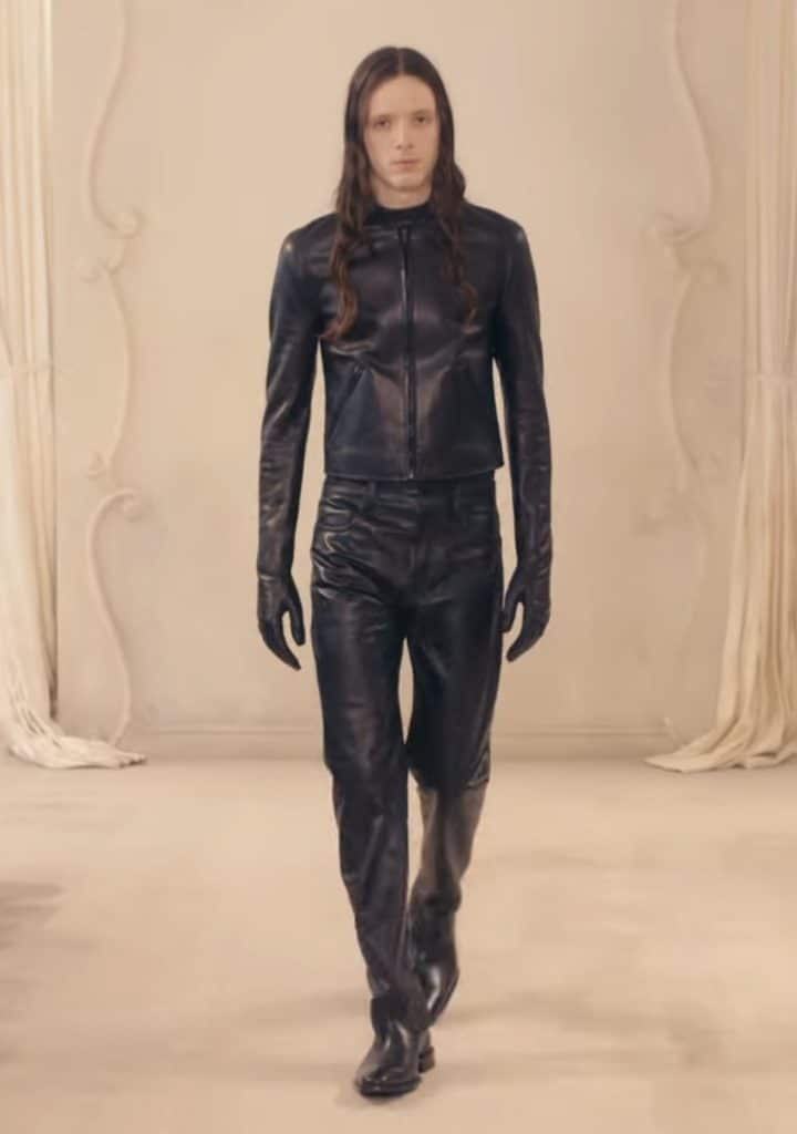 A model walks down the runway in a full leather outfit, similar to that of a biker outfit. He is also wearing leather gloves.