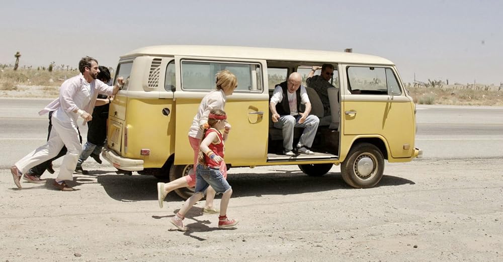 Two people push a yellow van as a mom and daughter run beside it.