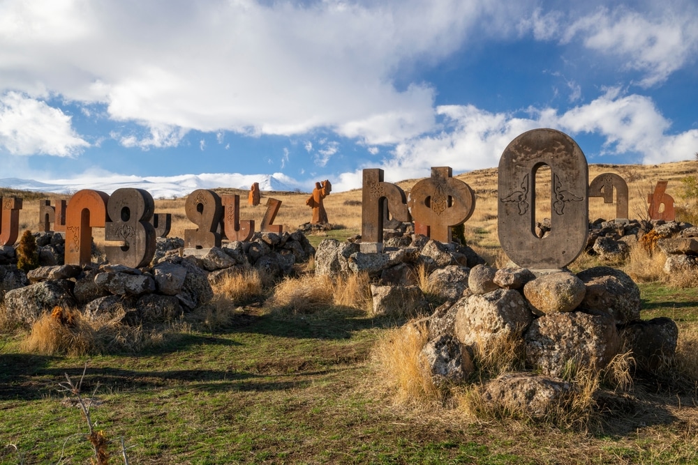 Stone sculptures of Armenian alphabet letters arranged outdoors on a grassy landscape with scattered rocks and grass, under a partly cloudy sky in Abaran, Armenia.