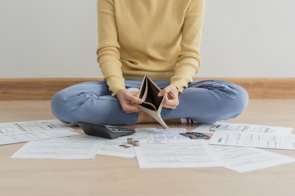 A feminine person holding an empty wallet and sitting on the floor. They are surrounded by bills, and spare change dots the papers.