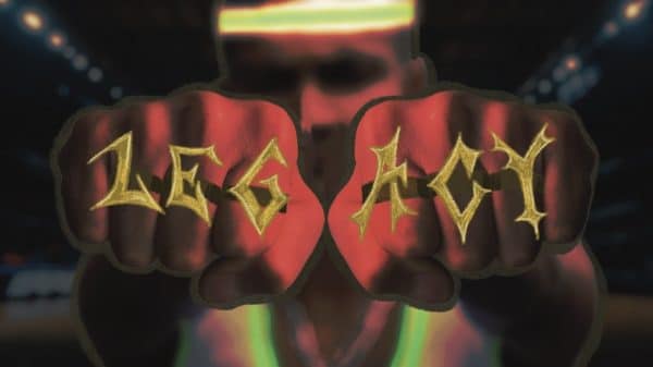 Close-up of two fists with the word ‘LEGACY’ tattooed across the knuckles in bold gold letters, with a blurred basketball court background and a player wearing a headband in the distance.
