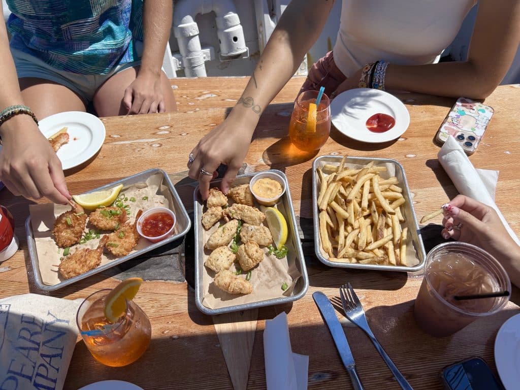 Three trays of food including coconut shrimp, haddock bites, and french fries paired with refreshing glasses of Aperol spritz at Pilot House.