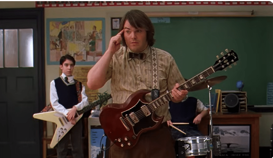 A man holding an electric guitar in a classroom. A student holding a guitar and a student playing drums are behind him.