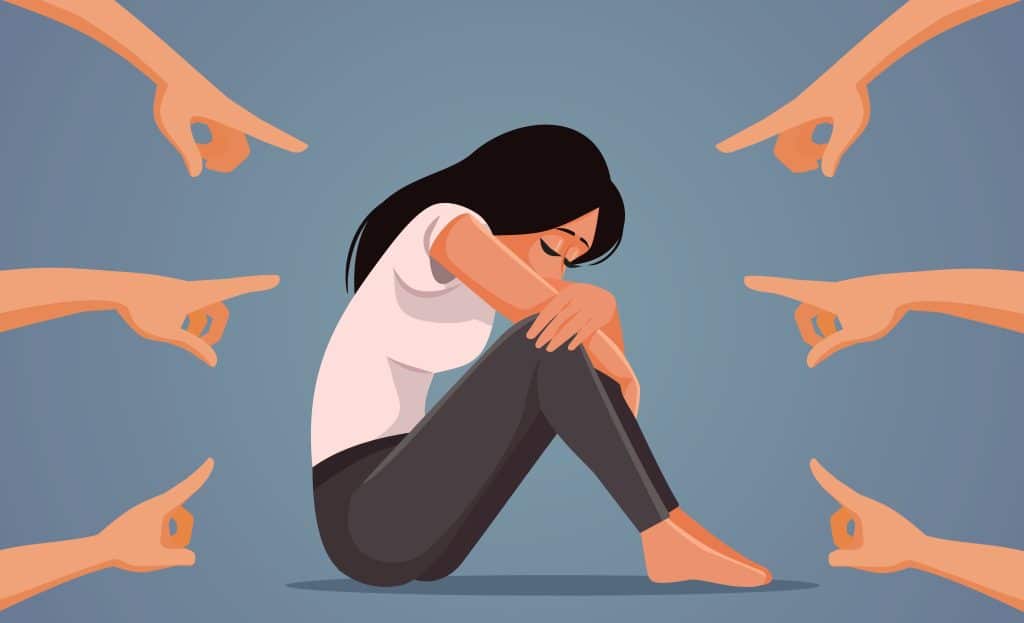 Young women in distress, siting with her arms wrapped around her knees, as people around her condemn her.