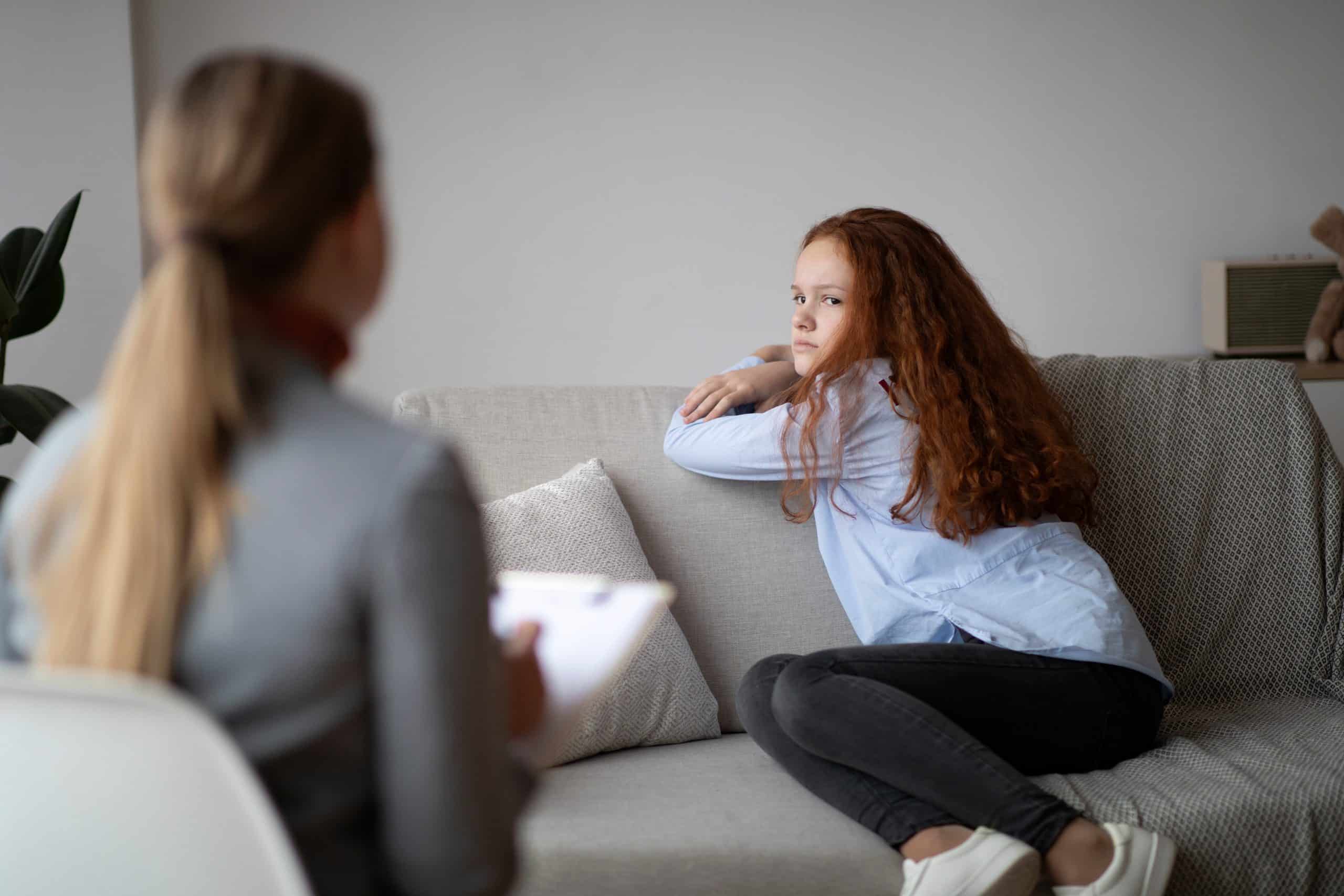 A woman sits on the sofa with a pained expression, while another woman looks at her.