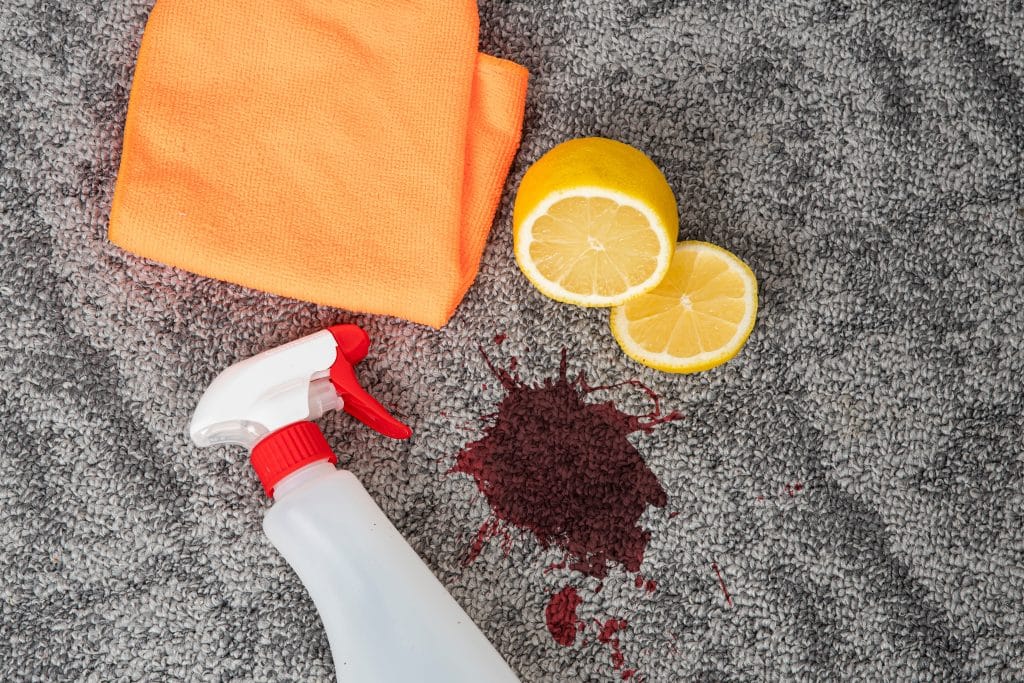 Spill on the carpet surrounded by cleaning bottle, rag, and lemons.
