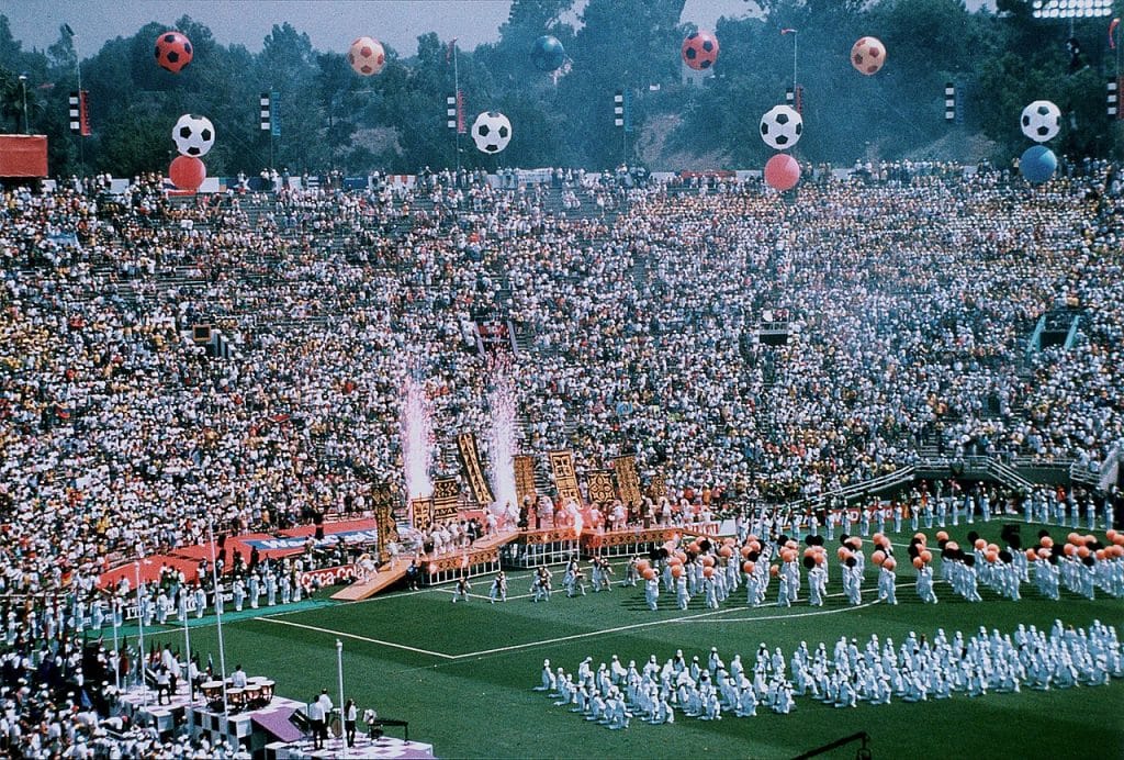 Pyrotechnics flare in front of a packed stadium. Two teams line up on a soccer pitch for the opening match of the World Cup in 1994.