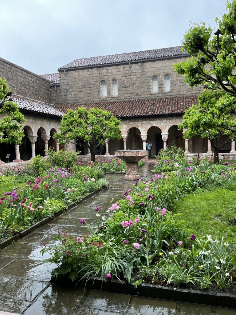 A view of a garden in the Met Cloisters full of flowers and trees with a path down the middle leading to arches in front of the museum building.
