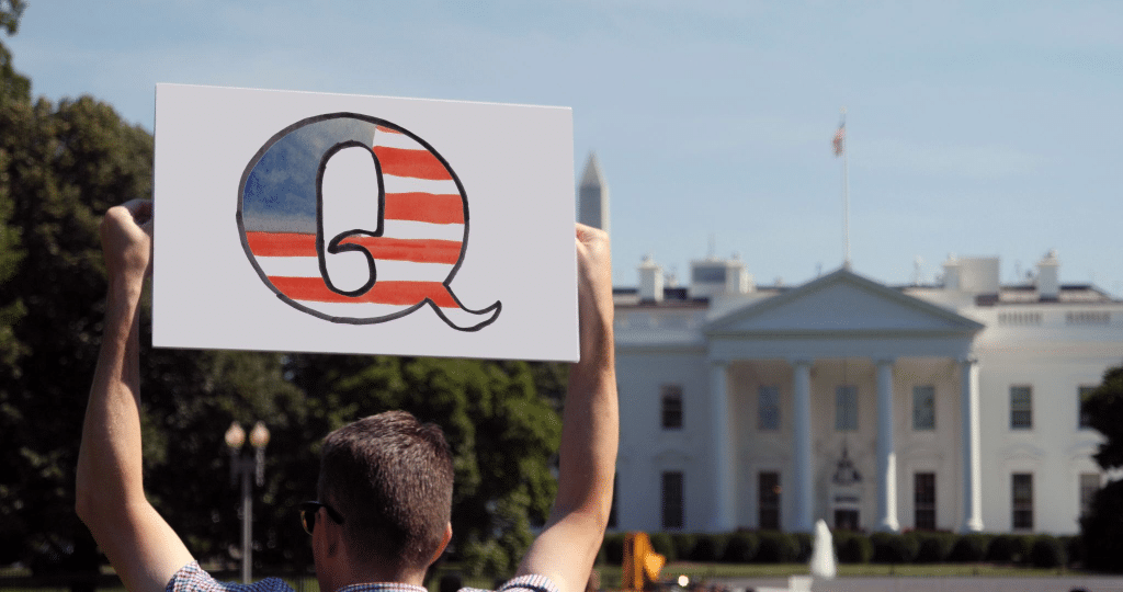 A man holds up a Qanon (the conspiracy theorist group) sign outside of the White House. The sign is a letter Q with the colors and design of the American flag.