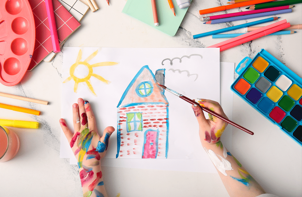 Child's hand drawing home with brush, watercolor paints and different stationery on white background