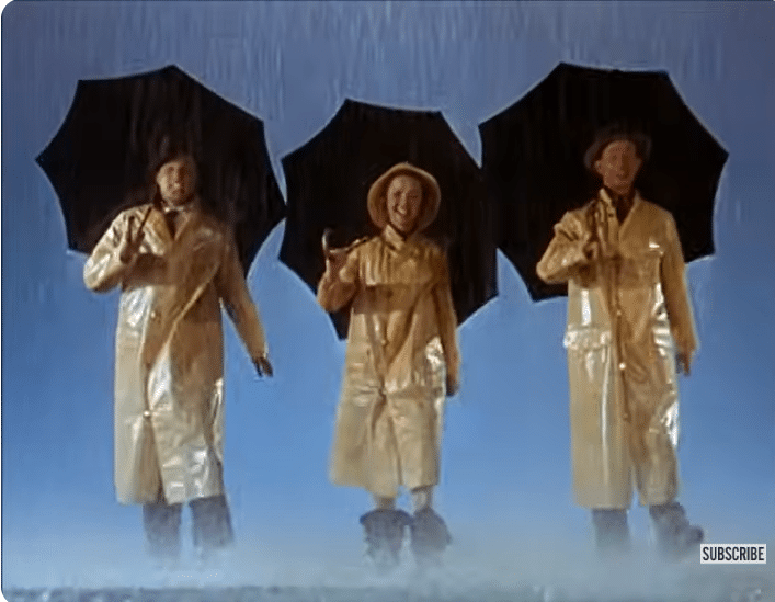 Two men and a woman wearing yellow raincoats and holding black umbrellas smile whilst it is raining heavily