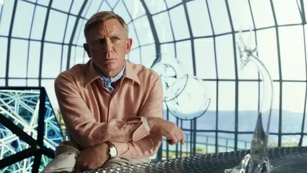A man in a pink shirt, arms crossed, surrounded by windows and glass sculptures.