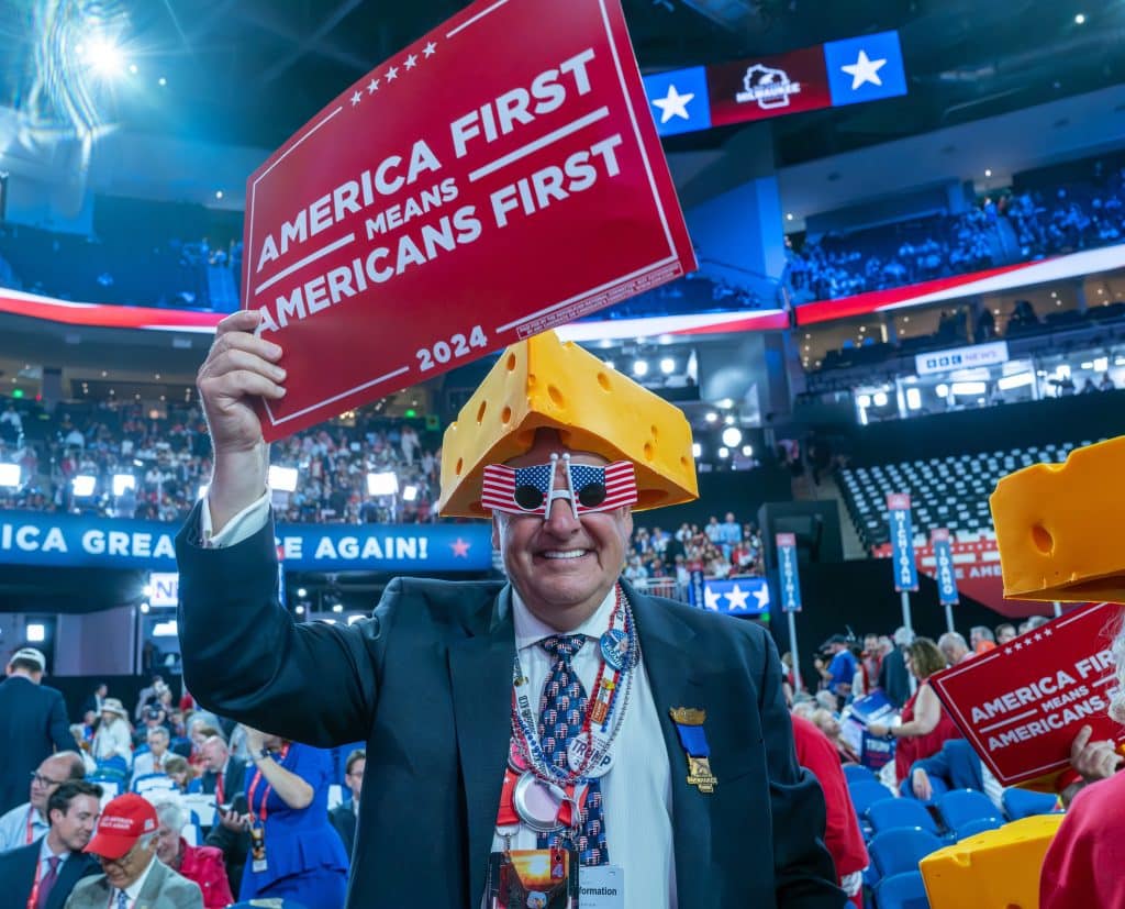 Attendant of the 2024 RNC holding a sign that reads "America First Means Americans First." The gentleman stands before a crowd in the half-packed stadium.