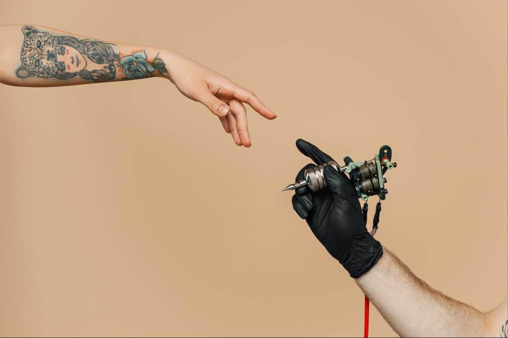 A tattooed arm reaching out to touch a gloved hand holding a tattoo machine, in a mirror of the 'Creation of Adam' painting.