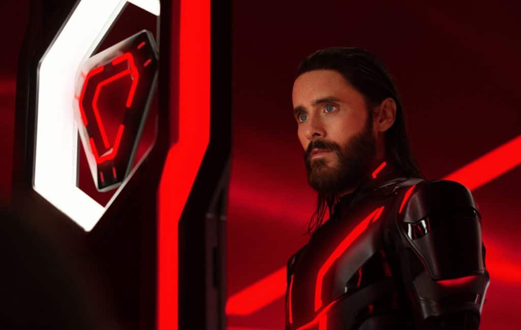 Jared Leto in black armor staring at a triangular red and black disc held in a shelf.