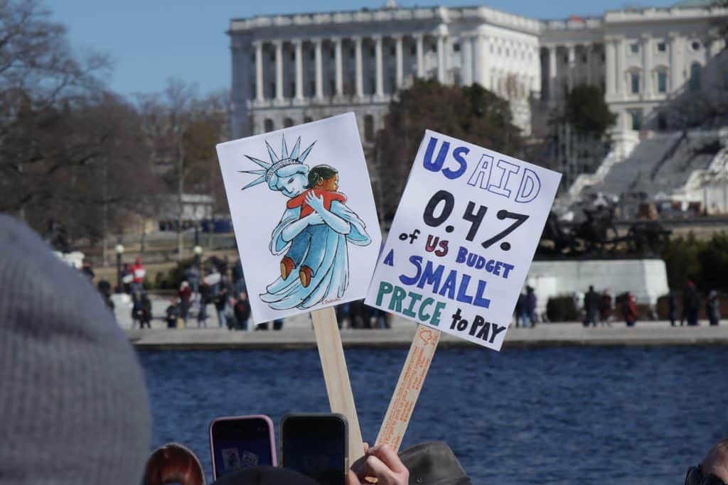 Protest signs held in front of the American capital. One reads "USAID 0.47 of US Budget. A small price to pay." The other depicts the Statue of Liberty embracing a young girl.