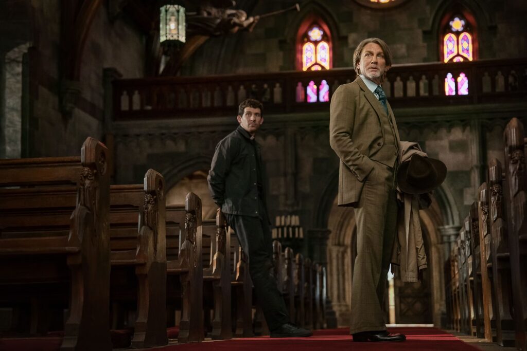 A man in a brown suit stands in a church, standing next to a priest in a black jacket.