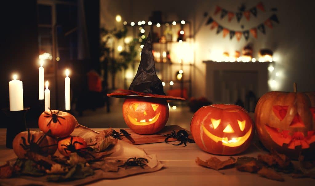 Halloween indoor scene: Pumpkin lanterns, candles of varying heights, orange jack-o'-lanterns, and spiders adorn the table, bathed in warm light.