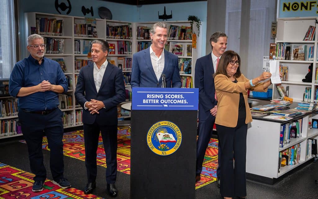 High Definition photo of Gavin Newsom giving a press release regarding the increased California Student test scores, there are notably smiles on the faces of ht e5 people in the photo including Newsom.