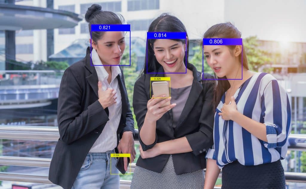 Three women smile at a phone, there are surveillance bounding boxes identifying each of their faces.