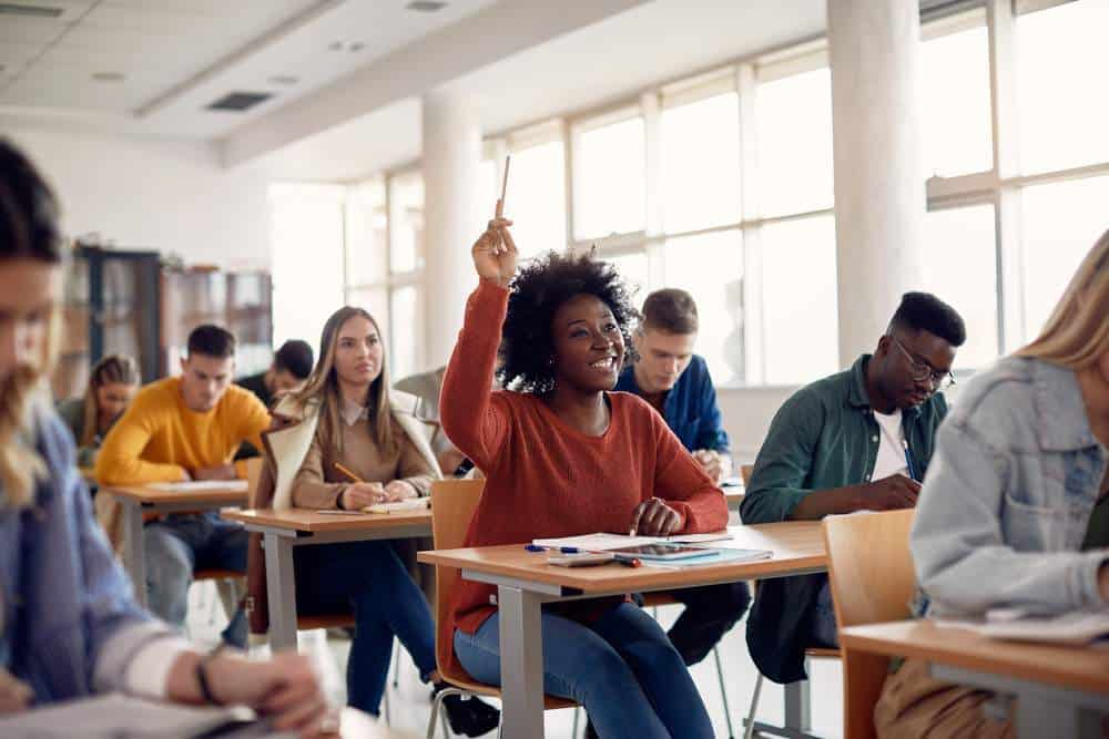 A college student smiling, sat at her desk with her hand raised, about to answer a question.