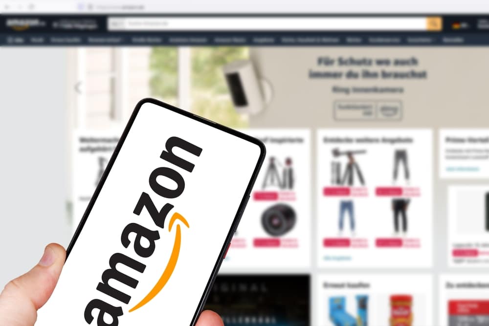 Someone holding a phone with the Amazon logo on it, with the website on a computer behind the phone, out of focus.