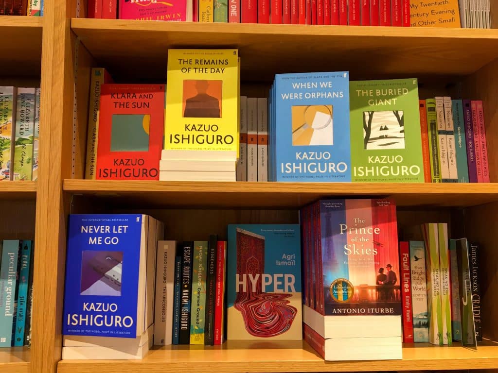 A series of books are facing forward on a shelf. They all read of different titles of Kazuo Ishiguro novels, displaying different collors and simple desings.