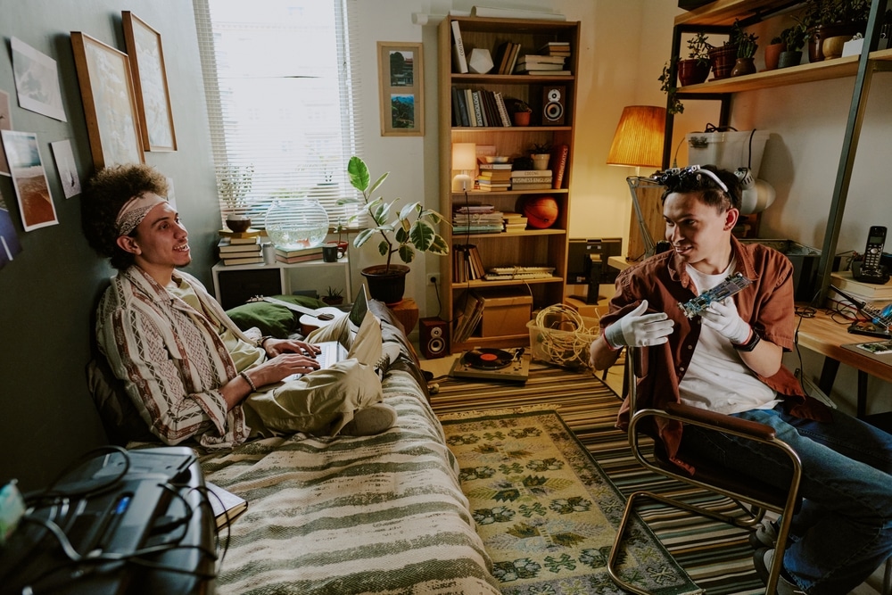 Two college boys sat in a dorm room. One is on a bed and one is on a chair, they are talking to each other.