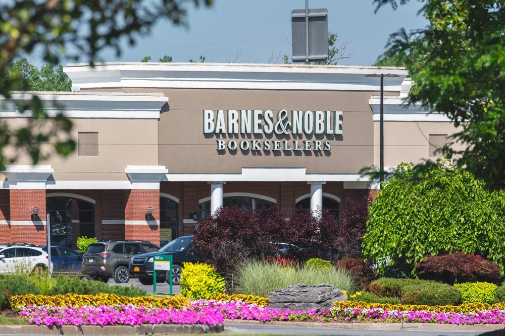 A storefront of a Barnes and Noble form across the street. There is foliage and purple flowers in the picture.