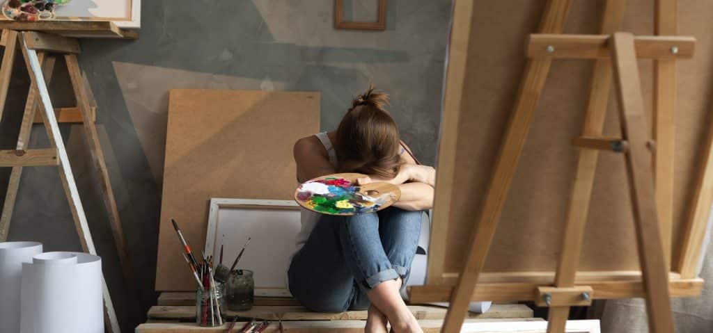 Image of a female artist in front of her painting canvas, holding her easel with her head resting on her knees, as though distressed.